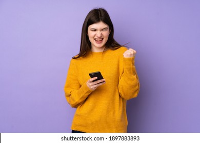 Young Ukrainian teenager girl over isolated purple background with phone in victory position