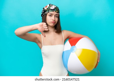 Young Ukrainian woman holding beach ball isolated on blue background showing thumb down with negative expression