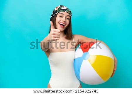 Young Ukrainian woman holding beach ball isolated on blue background with thumbs up because something good has happened
