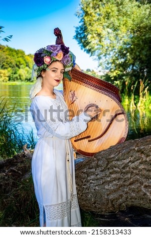 Young Ukrainian woman in national dress plays a bandura by the river on a summer sunny day.