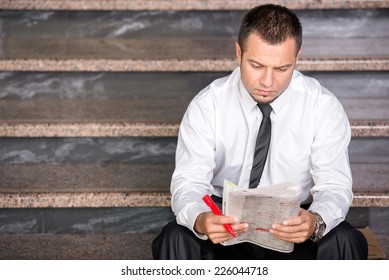 Young Unemployed Man Is Looking For A Job In The Newspaper, Sitting On The Stairs.