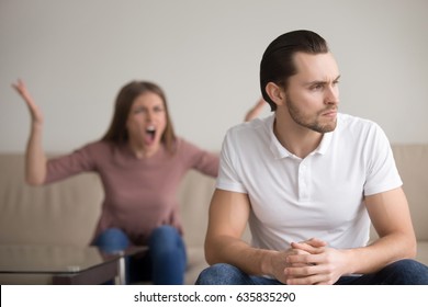 Young Unhappy Family Couple Quarrelling Sitting Apart At Home. Upset Frustrated Serious Husband Thinking About Problems Sitting In Foreground Looking Aside Ignoring His Hysteric Nagging Yelling Wife