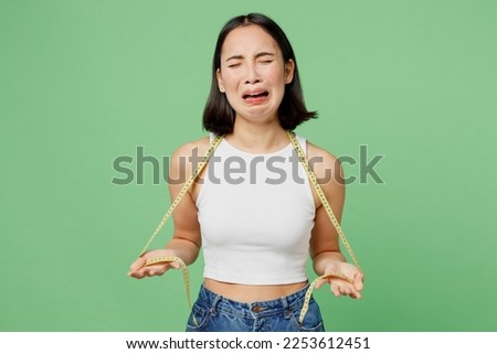 Young unhappy sad woman wearing white clothes hold measuring tape on neck crying isolated on plain pastel light green background. Proper nutrition healthy lifestyle fast food unhealthy choice concept