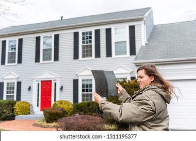 Young unhappy woman female homeowner standing in front of house on windy day in coat jacket during winter storm holding roof tile shingle inspecting damage