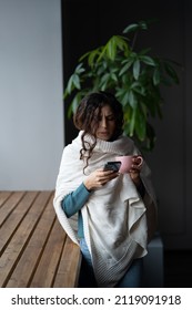 Young Unhealthy Woman Covered In Knitted Plaid Suffering From Cold Or Flu Writing Sick Day Email Message Via Smartphone While Standing Near Window At Home, Sick Female Letting Boss Know About Illness