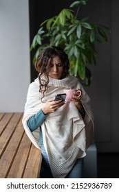 Young Unhealthy Woman Covered In Knitted Plaid Suffering From Cold Or Flu Writing Sick Day Email Message Via Smartphone While Standing Near Window At Home, Sick Female Letting Boss Know About Illness