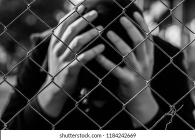 Young Unidentifiable Teenage Boy  Holding Hes Head At The Correctional Institute In Black And White, Conceptual Image Of Juvenile Delinquency, Focus On The Wired Fence.