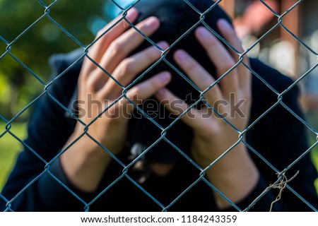 Young unidentifiable teenage boy  holding hes head at the correctional institute ,conceptual image of juvenile delinquency, focus on the wired fence.