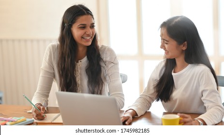 Young University Student Tutoring Their Lesson With Using A Computer Laptop While Sitting Together At Wood Working Desk Over Comfortable Sitting Room Windows As Background.