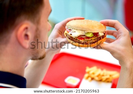 Young unrecognisable man, guy is sitting on food court in shopping mall, holding in hands juicy big burger, hamburger, French fries and soda, smiling. Fast junk unhealthy fat food. View from behind