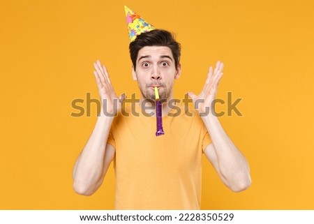 Young unshaved cute happy fun friendly caucasian man 20s in casual t-shirt wearing birthday hat cone blow pipe celebrating greeting spreading hands isolated on yellow color background studio portrait