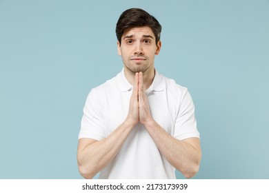 Young Unshaven Caucasian Man 20s In White Casual Basic T-shirt Hand Folded In Prayer Gesture, Begging About Something Isolated On Pastel Blue Color Background Studio Portrait. People Lifestyle Concept