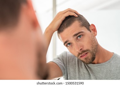 Young Unshaven Man Looking At Mirror In Bathroom At Home. Handsome Guy Looking At His Face In Mirror, Checking Hair And Hairline. Man In Pijamas Concerned With Hair Loss.