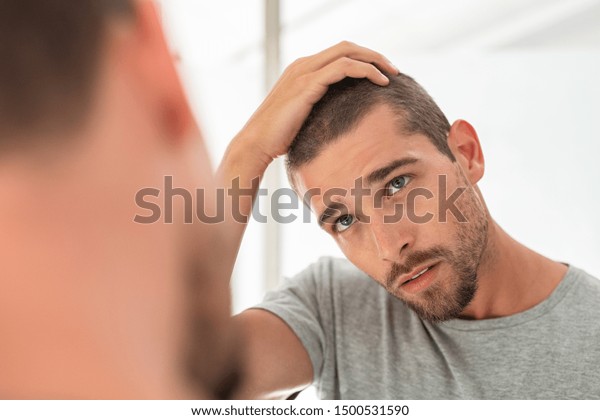 Young unshaven man\
looking at mirror in bathroom at home. Handsome guy looking at his\
face in mirror, checking hair and hairline. Man in pijamas\
concerned with hair loss.