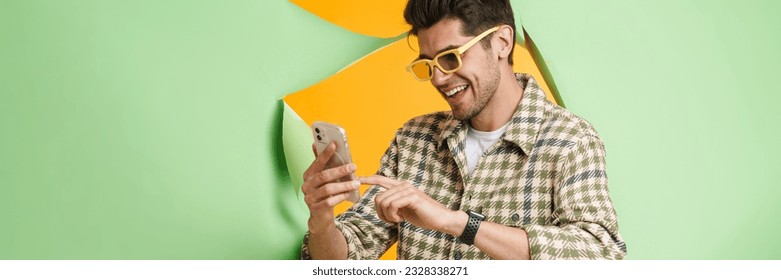 Young unshaven man smiling and using mobile phone while posing into hole in green background - Powered by Shutterstock