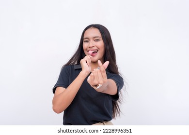 A young upbeat asian woman makes a finger heart sign while holding a lollipop. Isolated on a white background. - Shutterstock ID 2228996351