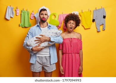 Young upset parents nurse baby, tired mother yawns and wants to sleep, father with diaper above head holds newborn son wrapped in blanket, isolated over yellow background, washed clothes on rope.