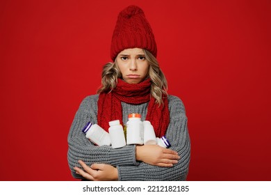Young upset sad woman wear grey sweater scarf hat hold many bottles with drugs pills isolated on plain red background studio portrait. Healthy lifestyle ill sick disease treatment cold season concept