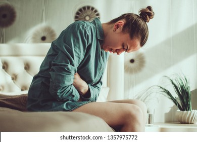 Young Upset Stressed Woman Suffering From Abdominal And Stomach Pain During Menstruation, PMS In Room At Home. Inflammation And Infection. Food Poisoning