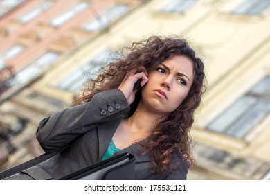 Young Upset Woman With Curly Dark Hair Dressed Business Style Talking On Phone Outdoor. Sad Girl. Business Problems. Failure In Business. To Get Upset. To Solve The Problem. A Phone Conversation.
