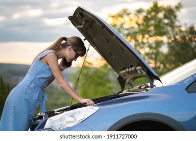 Young Upset Woman Driver Talking On Mobile Phone Near A Broken Car With Open Hood Inspecting Engine Having Trouble With Her Vehicle.