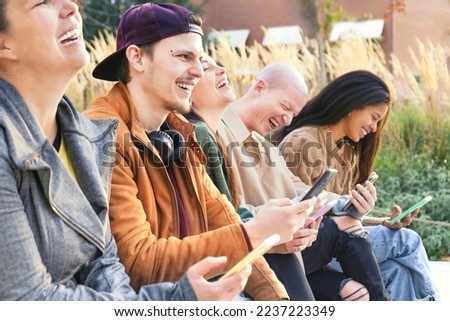 Young urban friends using smartphone outdoors sitting at park bench . Happy group of people laughing and sharing social content with mobile smart phone  at university college- Tech life style Concept
