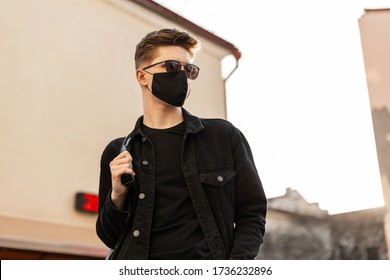 Young Urban Stylish Man Model In Fashion Black Denim Jacket With Backpack In Vintage Sunglasses In Black Trendy Protective Mask Poses In City On Bright Spring Sunny Day. Cool Guy Hipster In Street.