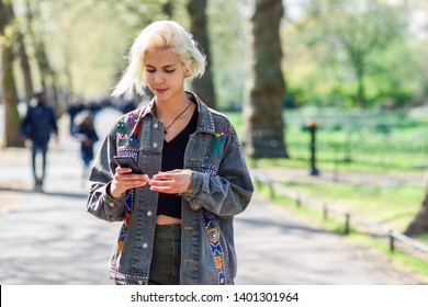 Young Urban Woman With Modern Hairstyle Using Smartphone Walking In Street In An Urban Park In London, UK.