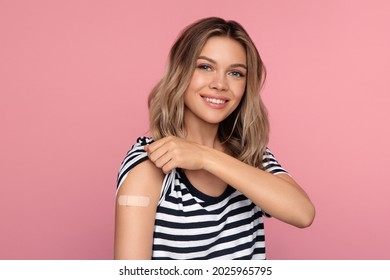 Young Vaccinated Woman Showing Arm With Adhesive Bandage After Vaccine Injection For Covid-19 Prevention With Happy Smile Isolated Over Pink Studio Wall. Coronavirus New Normal Reality And Vaccination