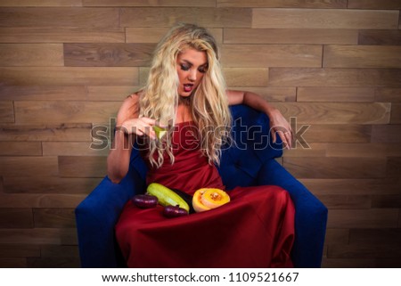 Young vegetarian woman sits with vegetables on armchair on background of wooden wall. Unusual housewife in elegant red dress holds vegetables and eats zucchini.
