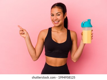 Young Venezuelan woman drinking a protein shake isolated on pink background smiling and pointing aside, showing something at blank space.