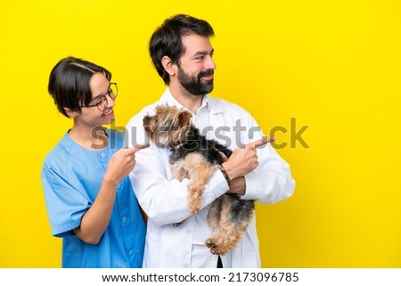 Young veterinarian couple with dog isolated on yellow background pointing to the side to present a product