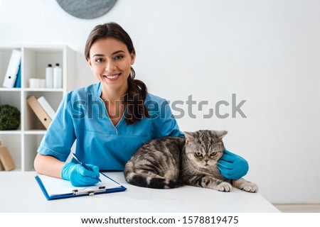 young veterinarian smiling at camera while writing on clipboard near tabby scottish straight cat