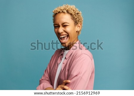 Young vibrant laughing blonde African female with nose ring winking keeping mouth opened widely, standing with arms folded against blue studio background with copy space for your advertisement