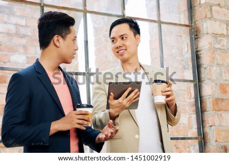 Young Vietnamese coworkers drinking take away coffee and discussing information on dgital tablet