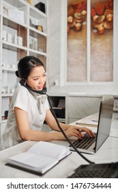 Young Vietnamese Female Spa Salon Owner Talking On Phone With Client And Working On Laptop