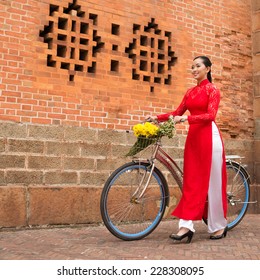 Young Vietnamese woman in traditional dress ao dai walking with a bicycle against a brick wall 