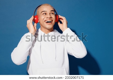 Young vivid happy smiling fun dyed blond man of African American ethnicity wear white hoody headphones listen music look aside on area isolated on plain dark royal navy blue background studio portrait