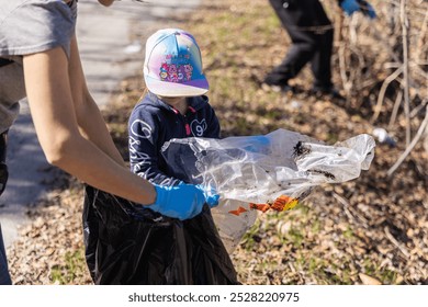 Young volunteer is putting trash in a garbage bag while her mother helps her during a community cleanup. Selective focus - Powered by Shutterstock