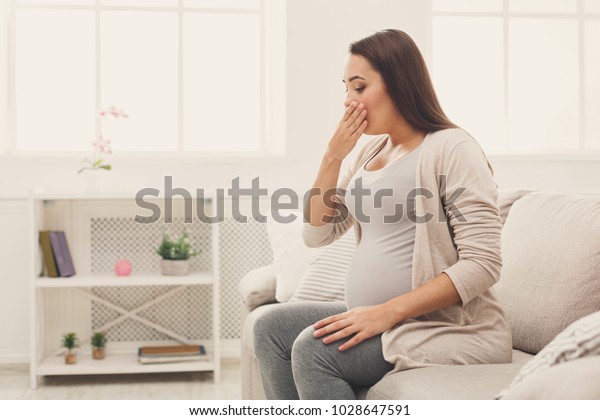 Young vomiting woman sitting on sofa. Pregnant lady\
suffering with nausea. Pregnancy symptoms, expectation, parenthood\
concept, copy space