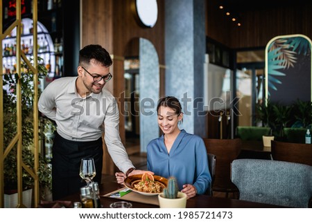 Young waiter with face protective mask serving delicious meal to beautiful woman in restaurant. 