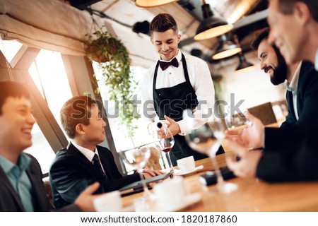 A young waiter pours wine into a glass to a Chinese businessman. Businessmen sit and chat in a restaurant while a waiter pours wine.