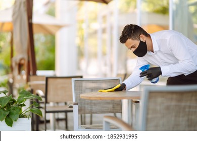 Young Waiter in protective face mask and gloves sanitizing surfaces, cleaning the table with disinfectant spray in a restaurant. Covid- 2019. - Powered by Shutterstock