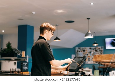 Young waiter serving customer at cash point in cafe. Man working with POS terminal. Cashier, barista checking for payment receipt. Hospitality, server and preparing a slip at the till in coffee shop