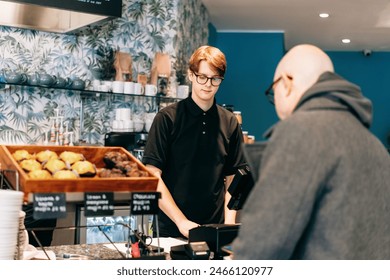 Young waiter serving customer at cash point in cafe. Man working with POS terminal. Cashier, barista checking for payment receipt. Hospitality, server and preparing a slip at the till in coffee shop. - Powered by Shutterstock