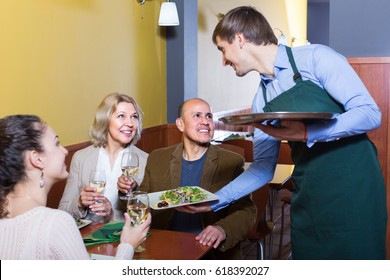 Young Waiter Serving Table In Restaurant With Happy Adults 
