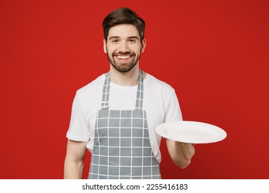 Young waiter smiling happy cheerful male housewife housekeeper chef cook baker man wear grey apron hold empty plate with workspace area isolated on plain red background studio. Cooking food concept - Powered by Shutterstock