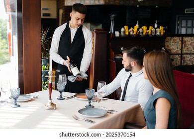 A young waiter in a stylish apron demonstrates and offers a fine wine to a beautiful couple in a restaurant. Customer service. - Powered by Shutterstock