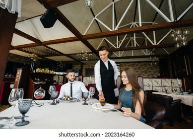 A Young Waiter In A Stylish Apron Serves A Table With A Beautiful Couple In A Refined Restaurant. Customer Service.