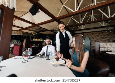 A Young Waiter In A Stylish Apron Serves A Table With A Beautiful Couple In A Refined Restaurant. Exquisite Delicacies Of Haute Cuisine Close-up.